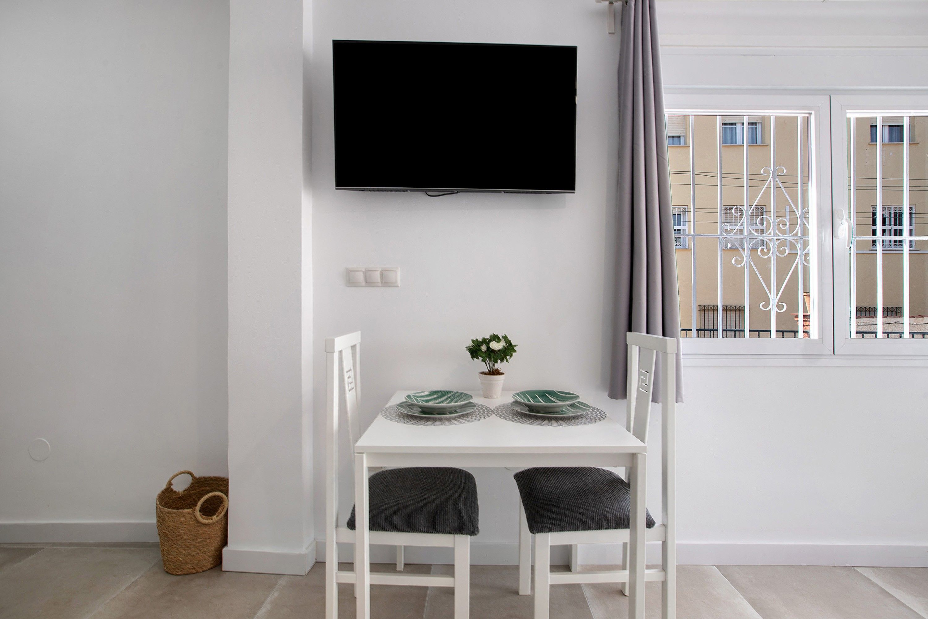 Ett modernt, minimalistiskt rum med en platt-TV som är monterad på väggen, ett litet bord med två stolar, en krukväxt och utsikt över en balkong med metallräcken.