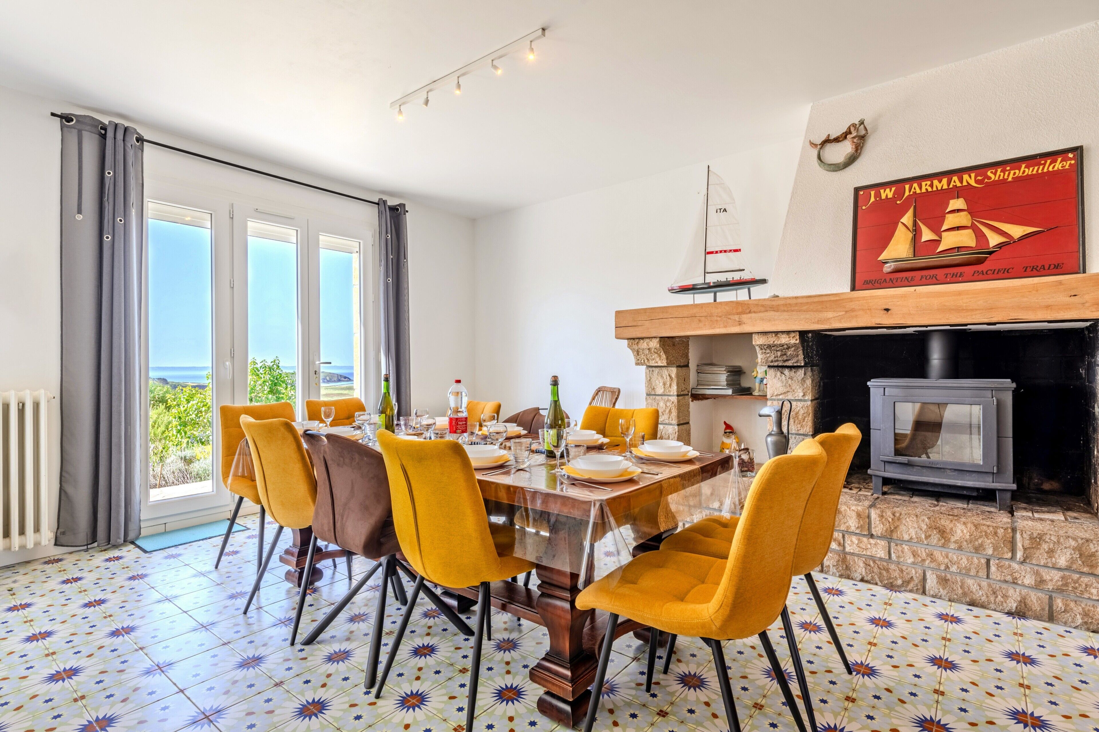 Un coin repas avec une table dressée pour quatre personnes, des chaises jaunes, une cheminée et une vue sur la mer.