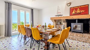 Uma área de refeições com uma mesa posta para quatro pessoas, cadeiras amarelas, uma lareira e vista para o mar.