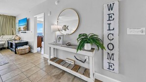 A modern living room with a white console table, a round mirror, a welcome sign, and a view of the ocean through a window.