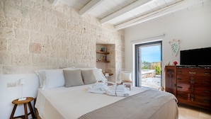 A bedroom with a stone wall, a bed with towels, a wooden nightstand, a TV, and a view through a door.