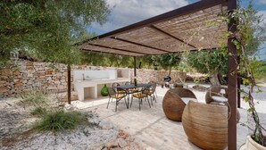 An outdoor patio with stone walls, a pergola, a dining table with chairs, and wicker furniture.