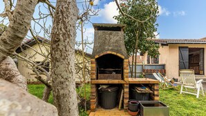 An outdoor brick oven with a chimney, surrounded by greenery and a white chair. - Holiday Home 'Blanche Neige Mimizan' with Private Terrace and Shared Garden (Mimizan)