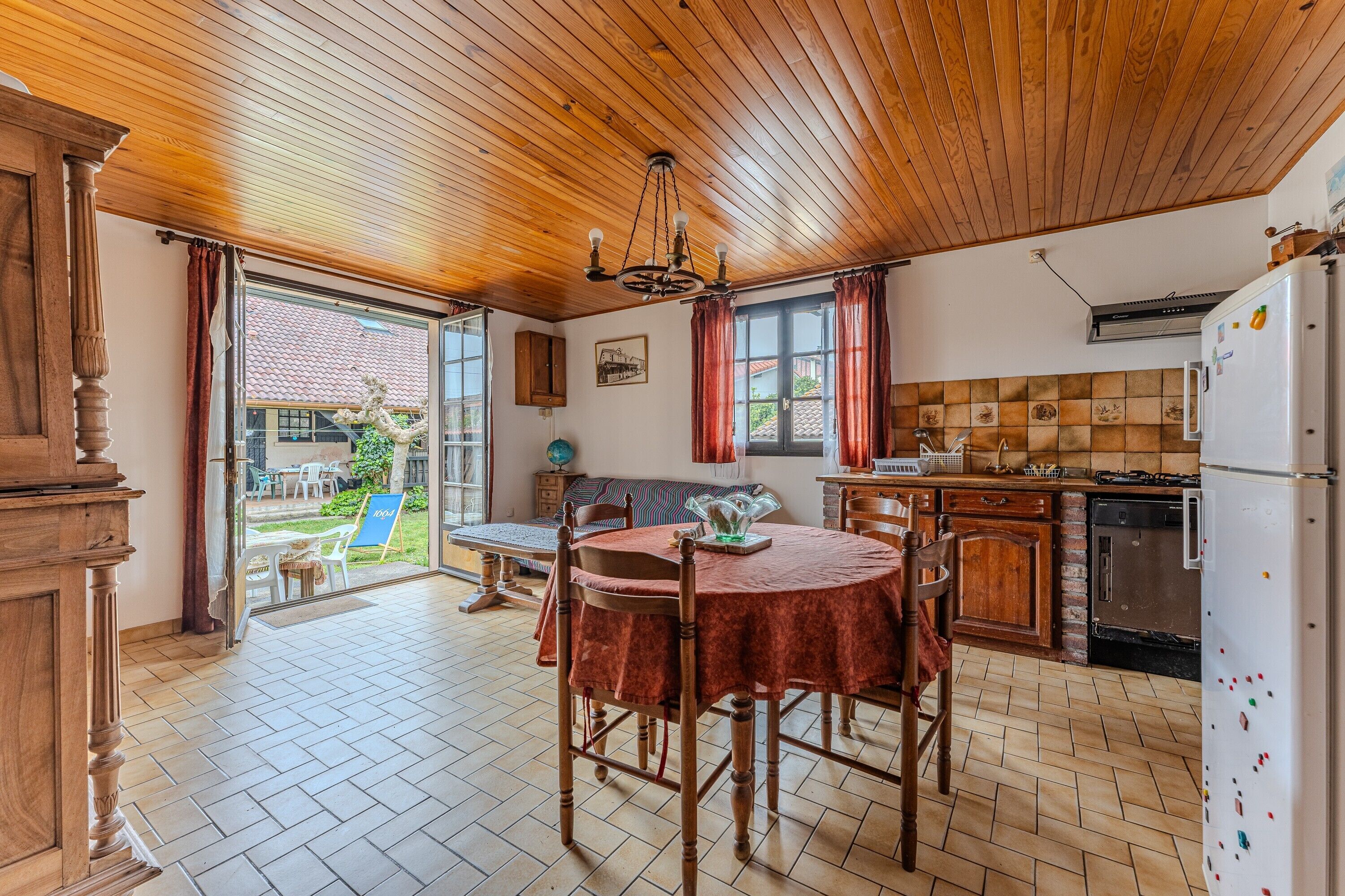 Une cuisine avec un plafond en bois, une table à manger avec des chaises, un réfrigérateur et une vue sur l’extérieur.