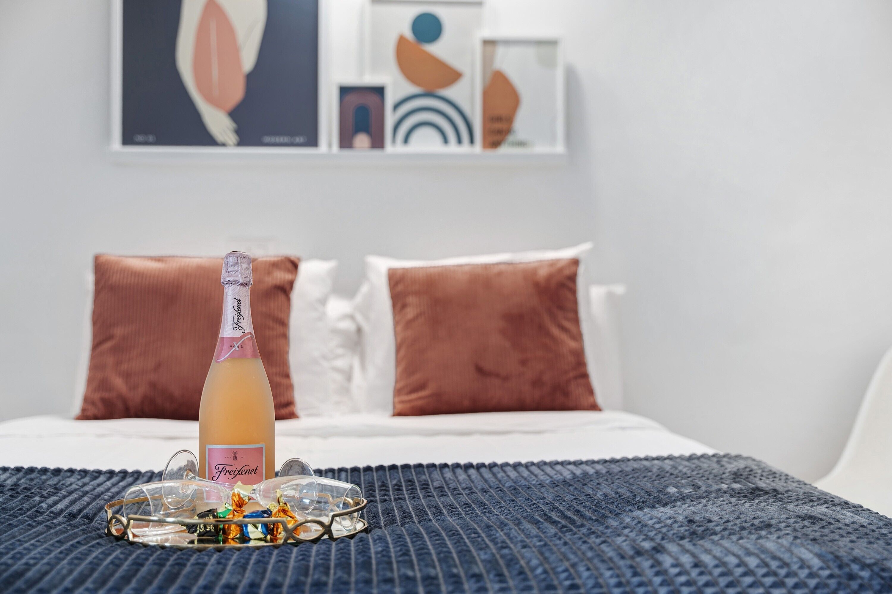 Una cama con una botella de Freixenet champagne, copas y bombones sobre un edredón azul con textura.