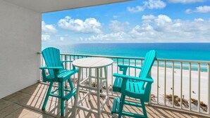 Un balcon avec deux chaises turquoise et une petite table, donnant sur une plage aux eaux d’un bleu limpide et au rivage sablonneux.