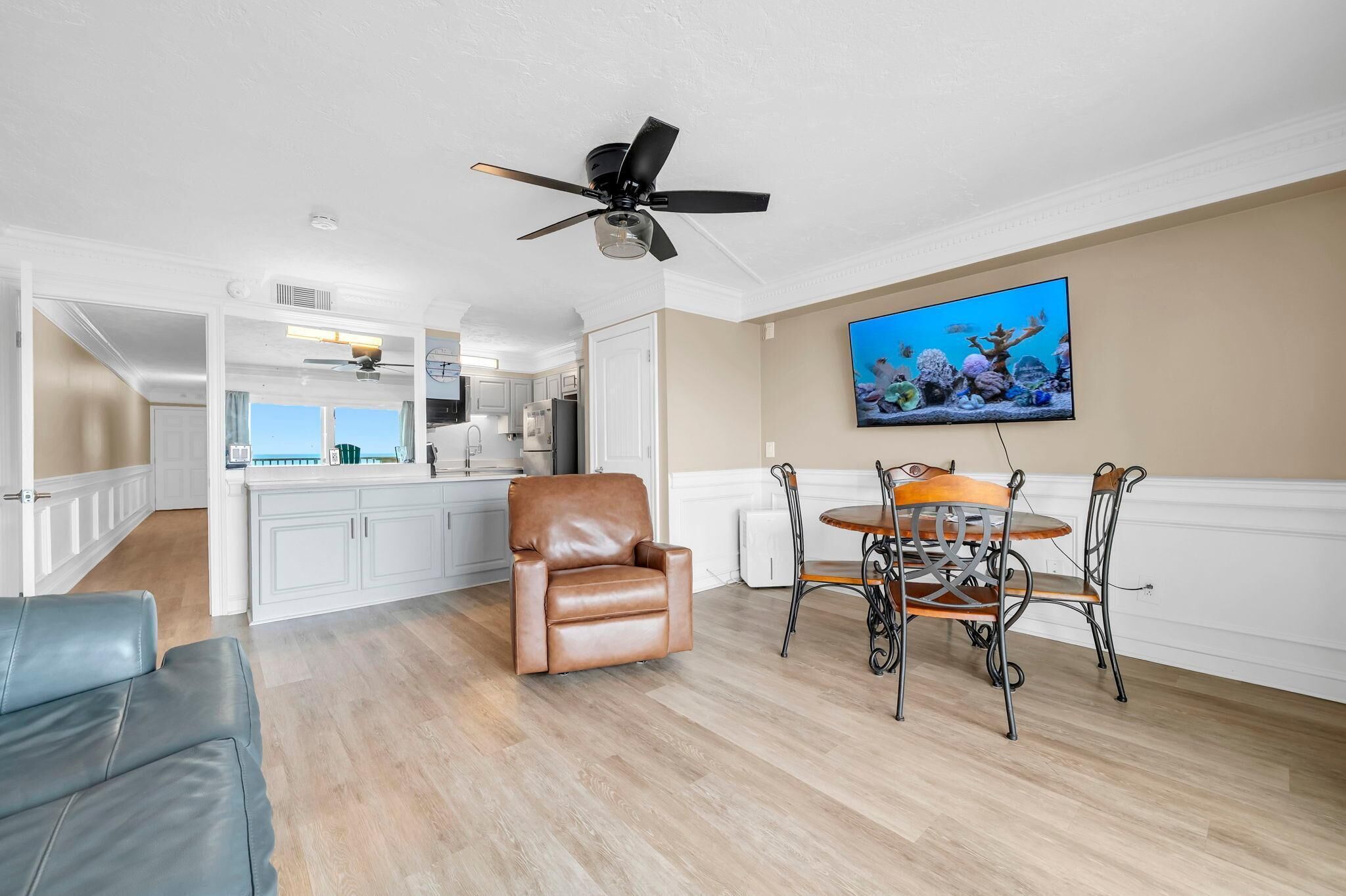 A spacious living room with a brown leather chair, a dining table with chairs, a ceiling fan, and a television mounted on the wall.
