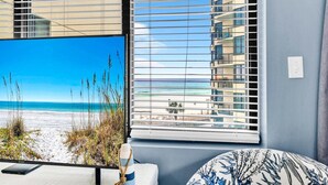 Ein Zimmer mit Blick auf den Strand durch ein Fenster, ein Bett mit einem gemusterten Kopfkissen, ein Nachttisch mit einer Lampe und eine wandmontierte Steckdose.