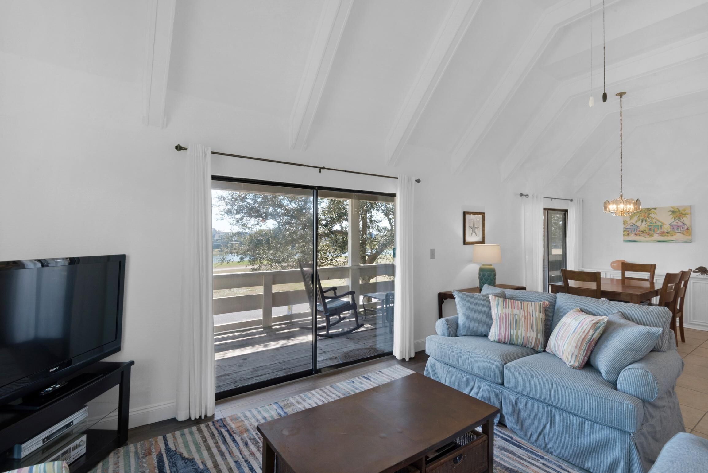 A spacious living room with a large TV, a blue sofa, a wooden coffee table, and a sliding glass door leading to an outdoor deck.