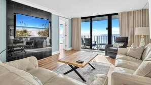 Ein modernes Wohnzimmer mit großem Fernseher, einem Holztisch und einer Couch. Ein Balkon bietet Blick auf den Strand.