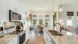 A modern kitchen with stainless steel appliances, a dining area with wicker chairs, and a large window providing natural light. - Shore Thing Getaway (Miramar Beach)