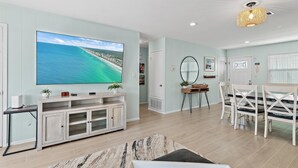 Una sala moderna con un televisor de pantalla plana grande, una zona de comedor con sillas blancas y una lámpara en el techo.
