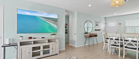 A modern living room with a large flat-screen TV, a dining area with white chairs, and a ceiling light fixture.