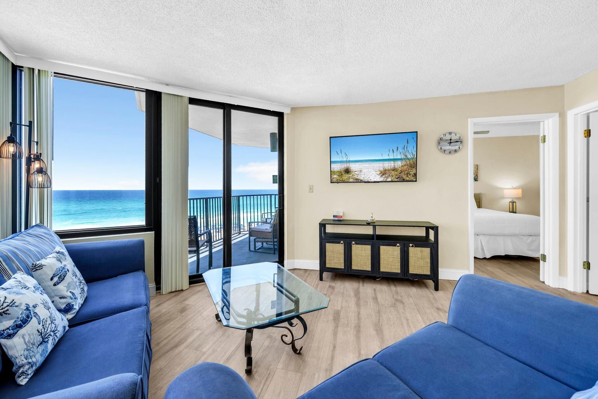 A room with a glass table, two blue sofas, a painting of a beach, a clock, and a view of the ocean.