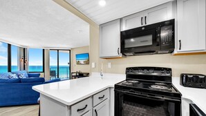 A modern kitchen with a stove, microwave, and toaster, a white countertop, and a view of the ocean. - Sunbird 508e (Panama City Beach)