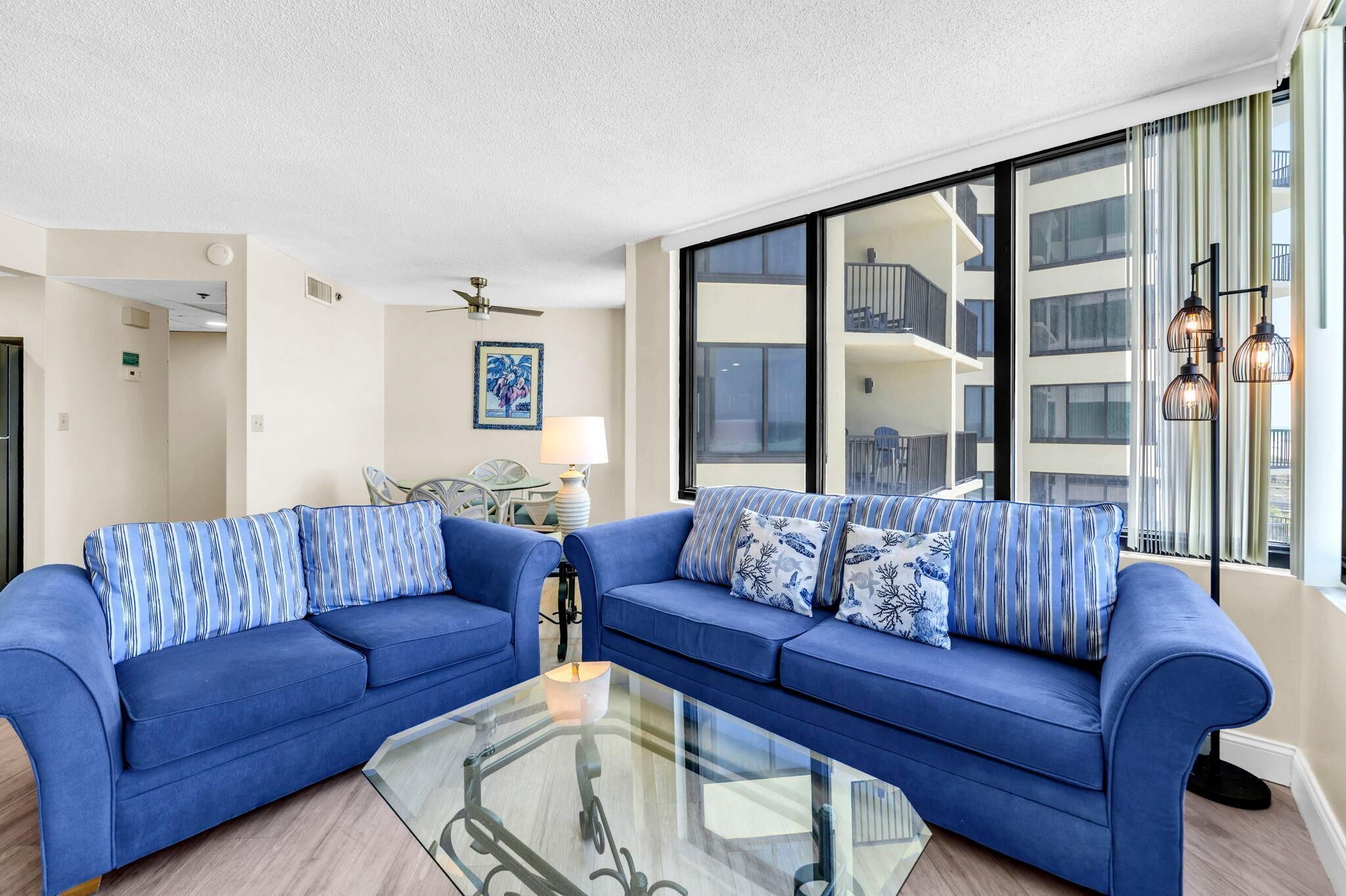 Ein modernes Wohnzimmer mit zwei blauen Sofas, einem Glastisch, einem Deckenventilator, einer Lampe und einem großen Fenster mit Blick ins Innere des Gebäudes.