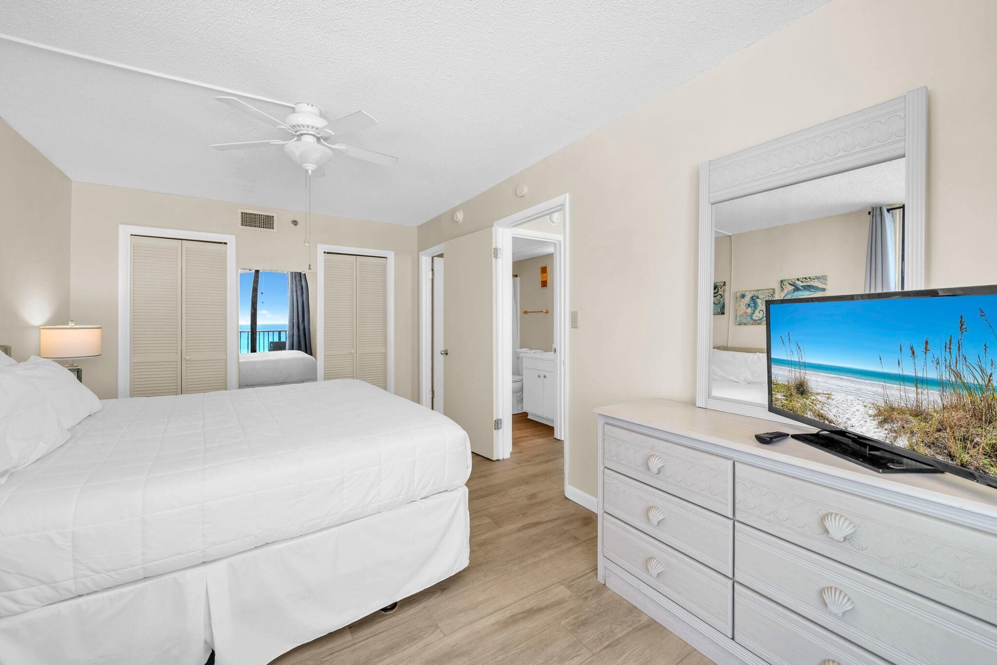 A bedroom with a bed, a ceiling fan, a TV on a dresser, and a view of the beach through an open door.