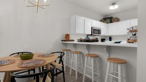 A modern kitchen with white cabinets, a wooden dining table with woven placemats, and bar stools. - Heron's Watch Way 4104- Coach's Sand Dollar (Santa Rosa Beach)