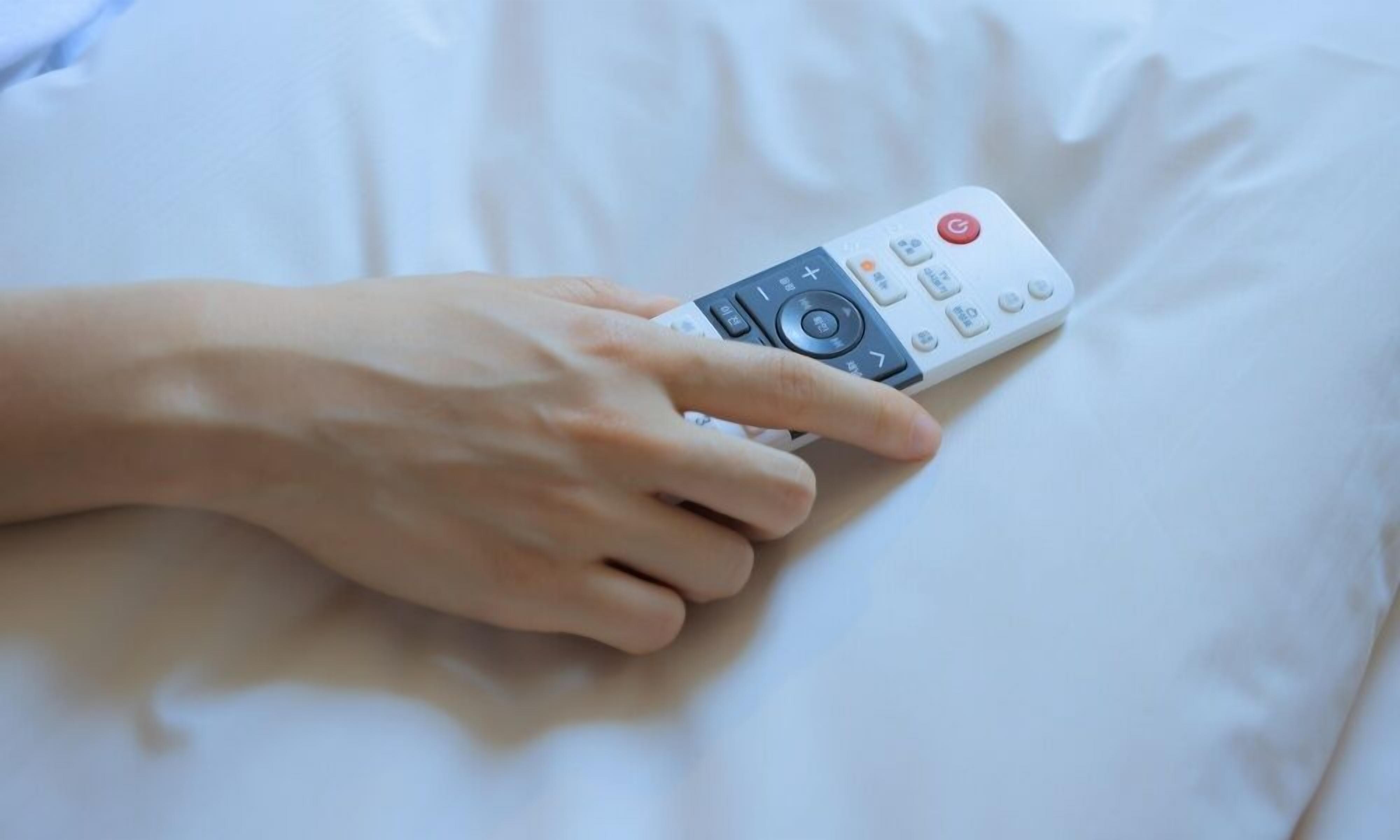 Una mano sostiene un control remoto sobre una cama blanca.