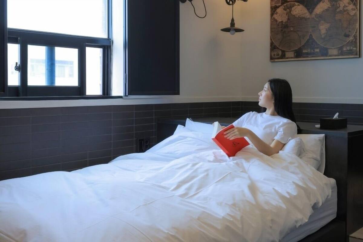 A modern bedroom with a bed, a nightstand, a window, and a wall-mounted light fixture.