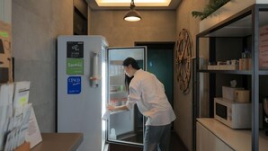 Eine Person in einem weißen Kittel öffnet einen Kühlschrank in einem schmalen Flur. An der Wand hängt eine Uhr, und auf der rechten Seite befindet sich ein Regal mit verschiedenen Gegenständen.