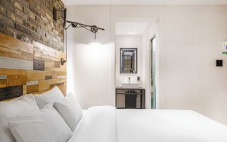 Habitación de hotel con pared de ladrillo, cama con ropa de cama blanca, lámpara colgante y vista al baño.