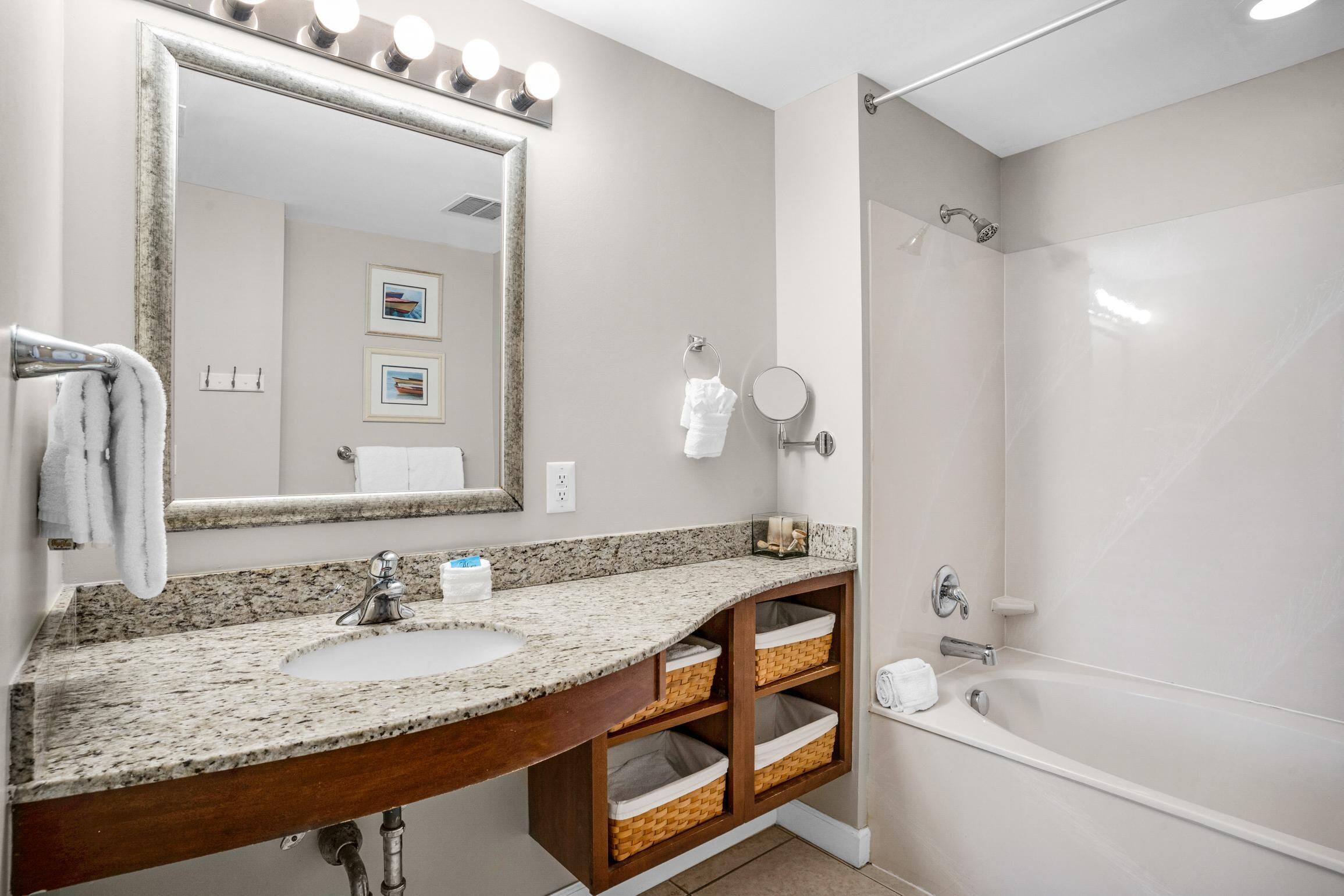 A bathroom with a granite countertop sink, a large mirror with lights, a shower, and wicker baskets under the sink.