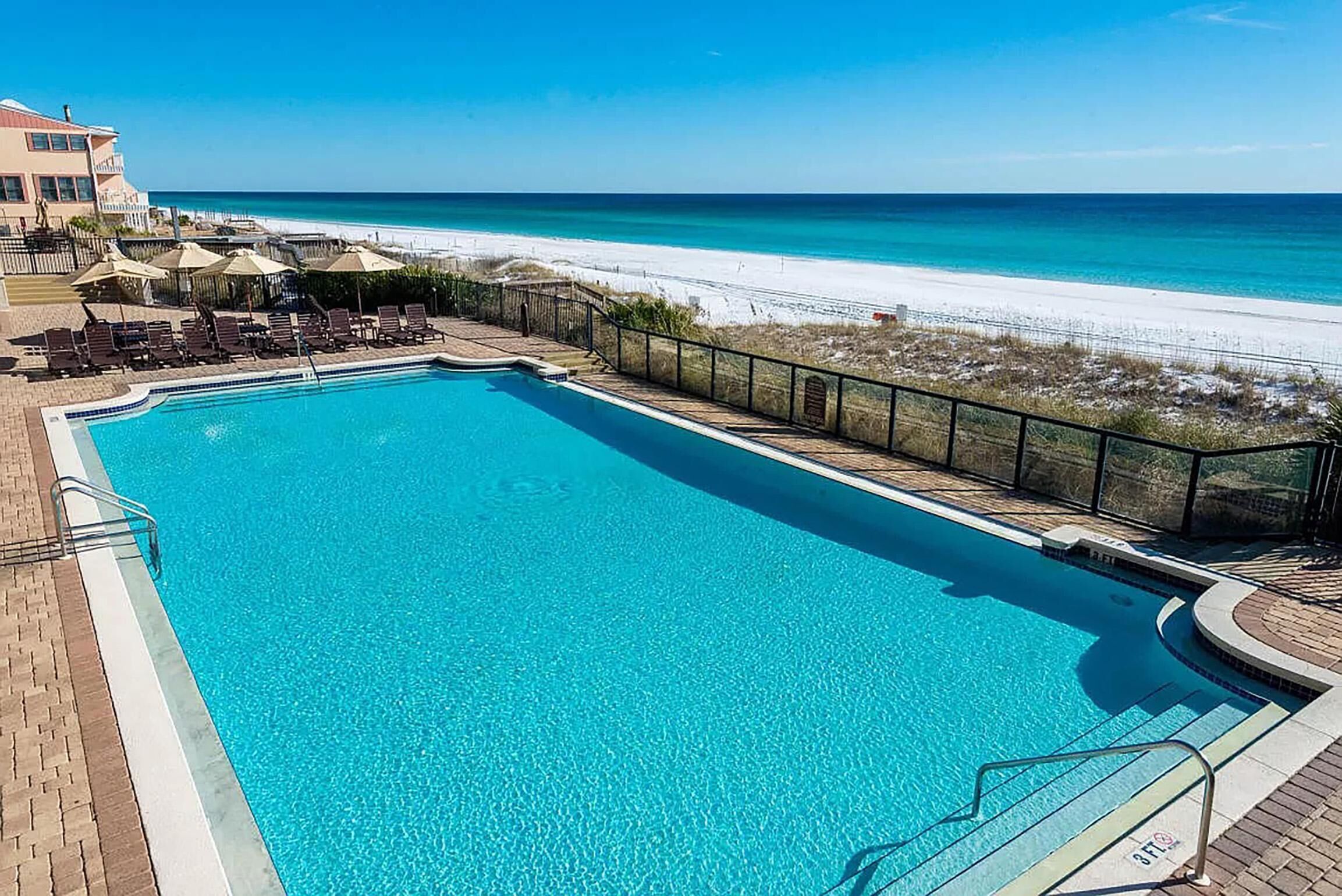 A clear blue pool with lounge chairs and umbrellas, a beach view, and a fenced area.