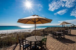 A beachside patio with tables and chairs, a large umbrella, and a view of the ocean.