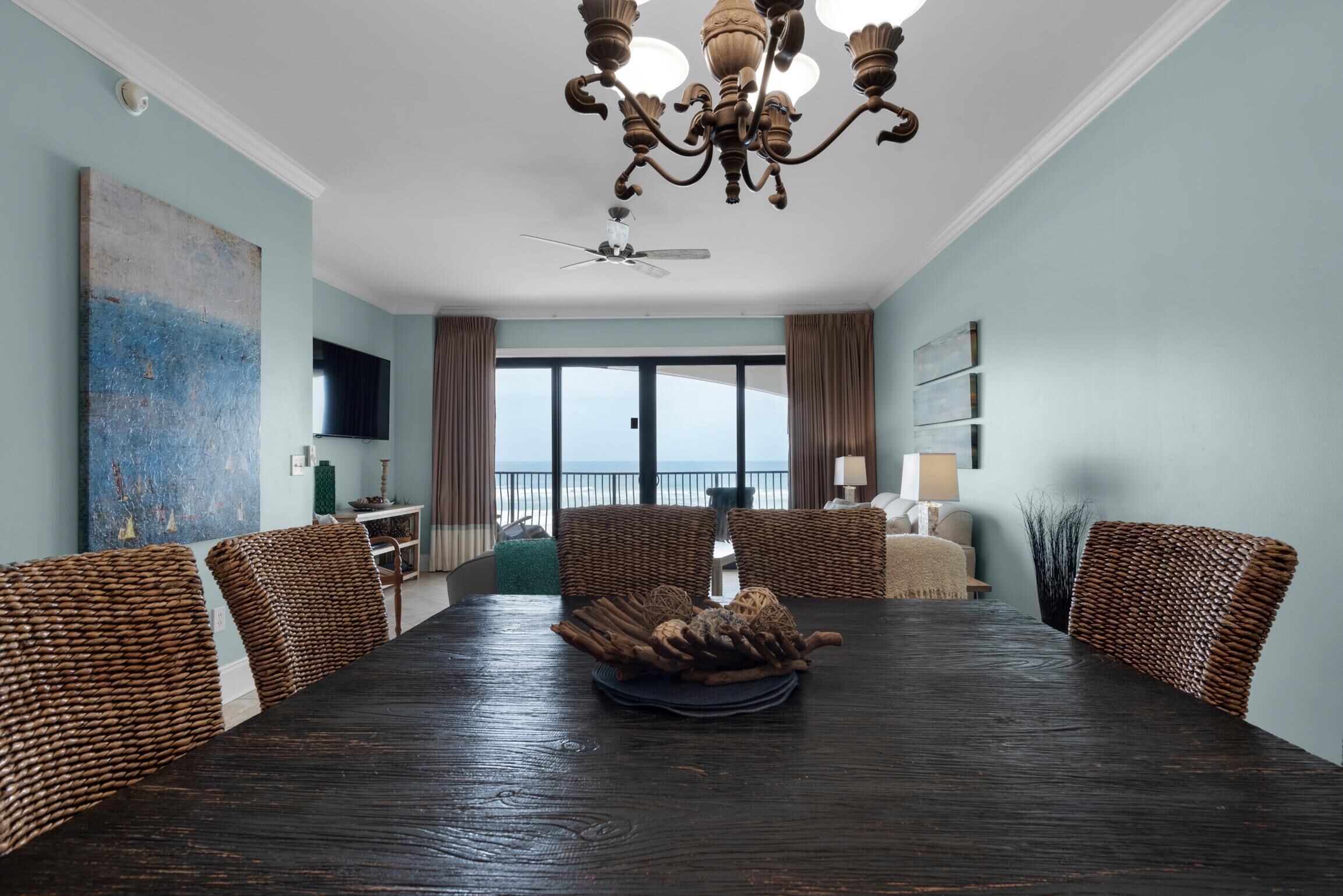 A dining room with a dark wooden table, wicker chairs, a chandelier, and a view of the sea through large windows.