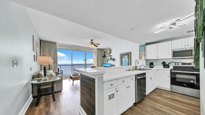 A modern kitchen with white cabinetry, a dishwasher, a stove, and a microwave. There is a dining area with a table and chairs, and a balcony with a view of the ocean. - Majestic Beach Towers 1-1911 (Panama City Beach)