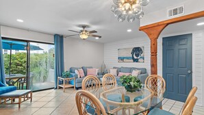A modern living room with a glass dining table, wooden chairs, a blue sofa, and a ceiling fan. - Southbay by the Gulf #122 - Salty Paws (Destin)