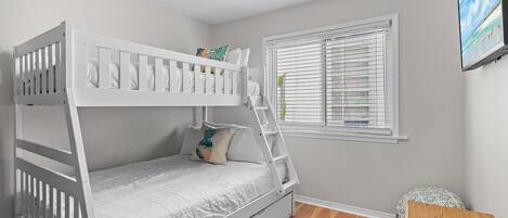 A bunk bed with a ladder, a small table with a remote control, and a window with blinds.