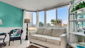 A living room with a beige sofa, a wicker chair, a floor lamp, and a view of palm trees through large windows.