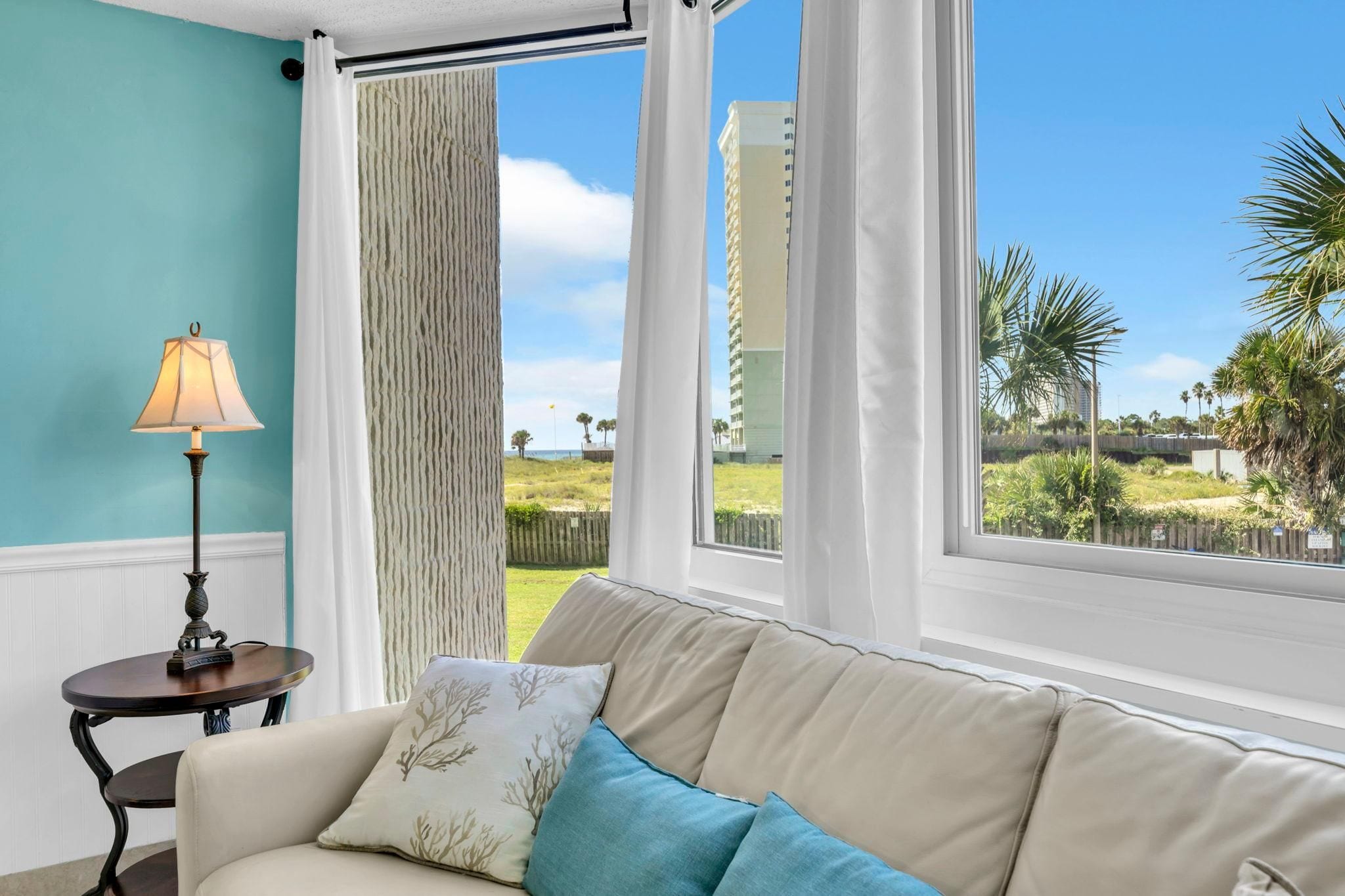 A room with a sofa, a lamp, and a view of a beach and buildings.