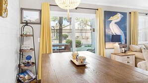 A dining area with a wooden table, yellow curtains, a chandelier, and a painting of a pelican. - Gulf Highlands 116h (Panama City Beach)