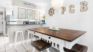 A kitchen with a wooden island, white bar stools, a chandelier, and decorative wall art including a sign that reads "OUR HAPPY PLACE" and letters "P C B". - Gulf Highlands 116h (Panama City Beach)