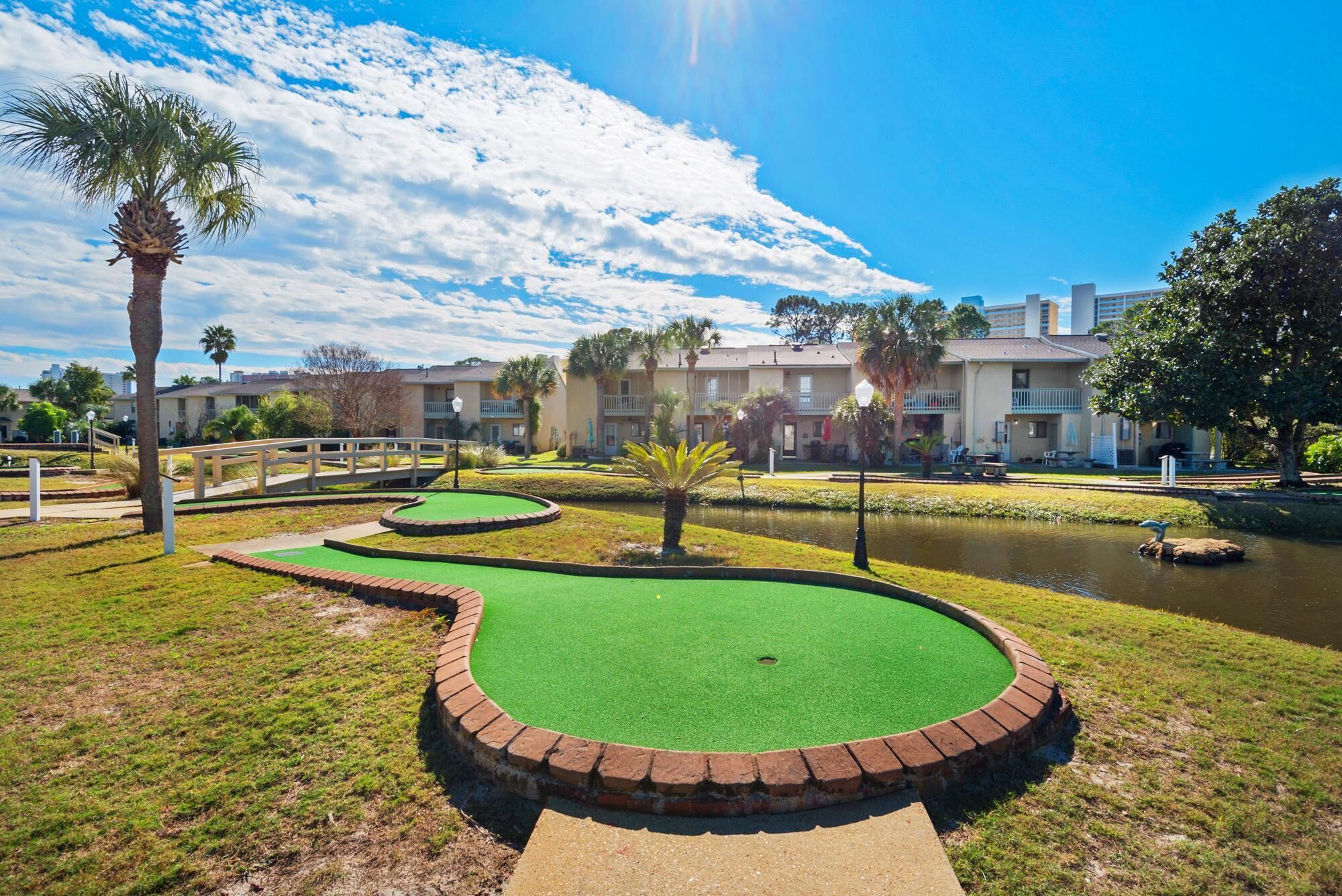 A golf course with a pond, palm trees, and a building with balconies in the background.