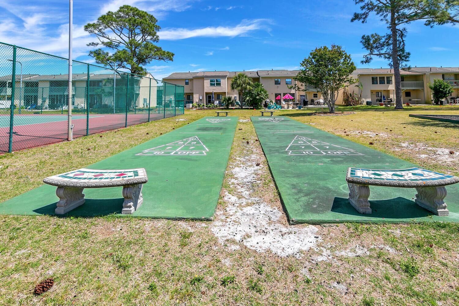 A shuffleboard court with two benches, a fenced tennis court, and a building in the background.