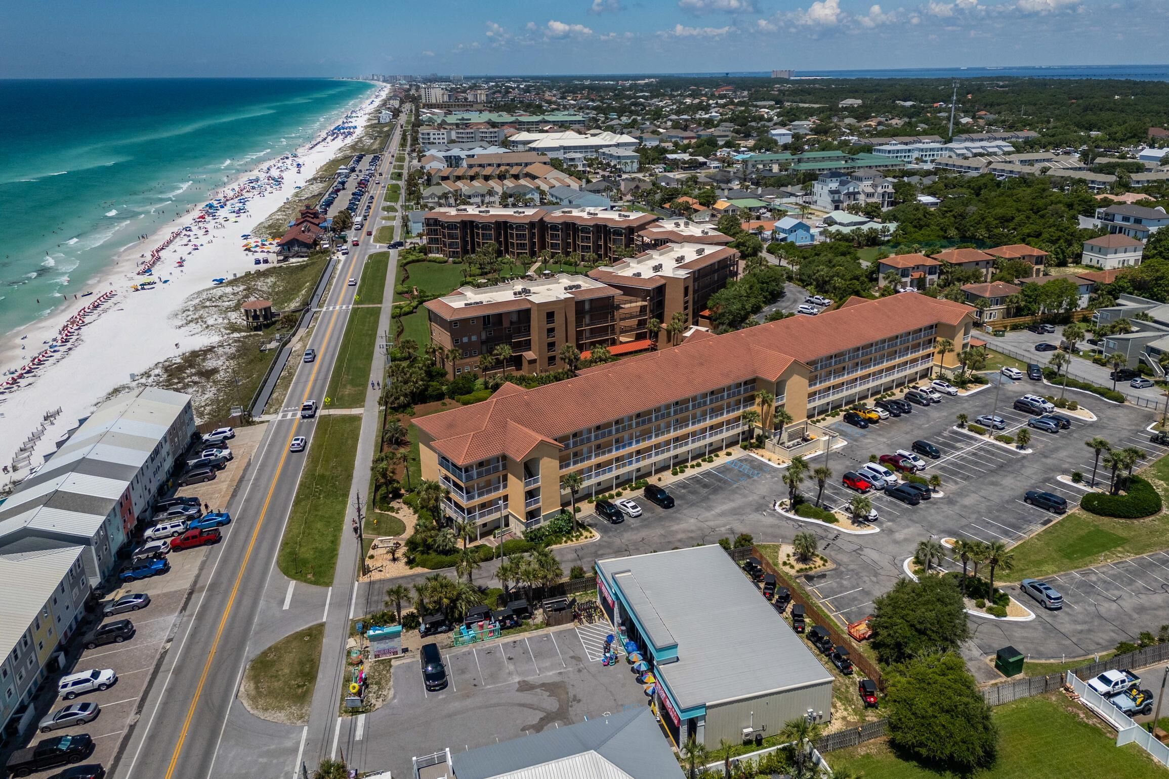 An aerial view of a coastal area with a beach, a parking lot, and several buildings.