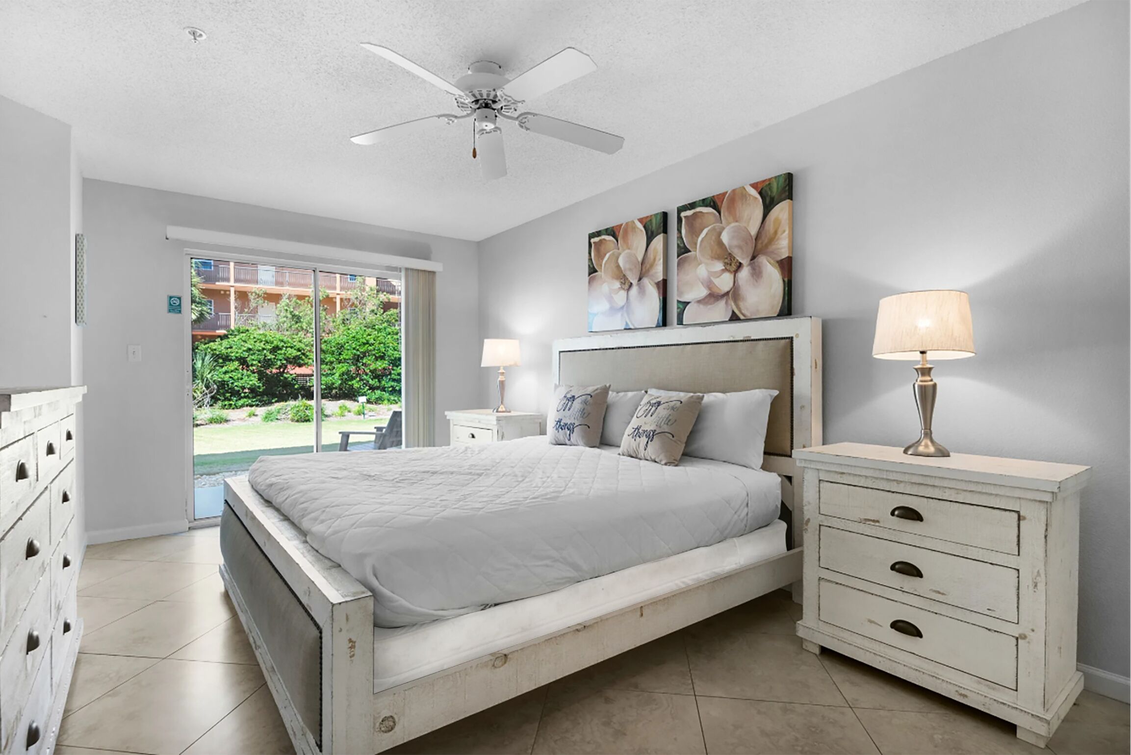 A bedroom with a bed, a nightstand, a dresser, a ceiling fan, and a window with a view of greenery outside.