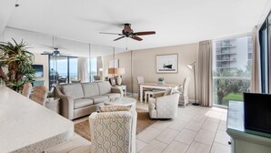 A modern living room with a sofa, armchairs, a coffee table, and a TV. There is a ceiling fan, a dining table with chairs, and a large window with a view of the outside. - Mainsail 213 (Miramar Beach)