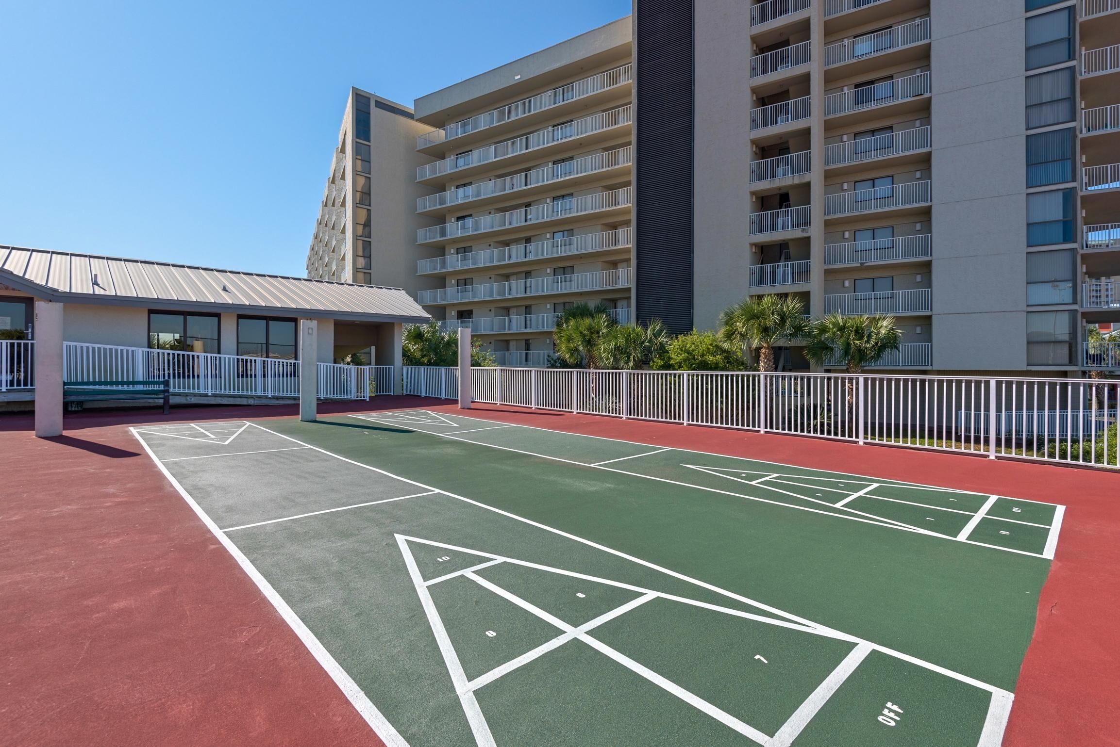 A courtyard with a shuffleboard court, a building with multiple balconies, and a fenced area with palm trees.
