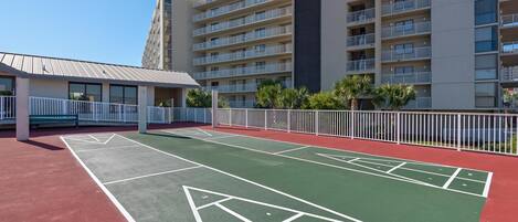 A courtyard with a shuffleboard court, a building with multiple balconies, and a fenced area with palm trees.