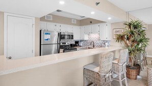 A modern kitchen with white cabinets, stainless steel appliances, and a central island with wicker chairs. - Mainsail 213 (Miramar Beach)