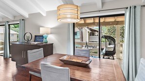 Un espace repas comprenant une table en bois, des chaises, un luminaire suspendu et une vue sur une terrasse extérieure, accessible par de grandes portes vitrées.