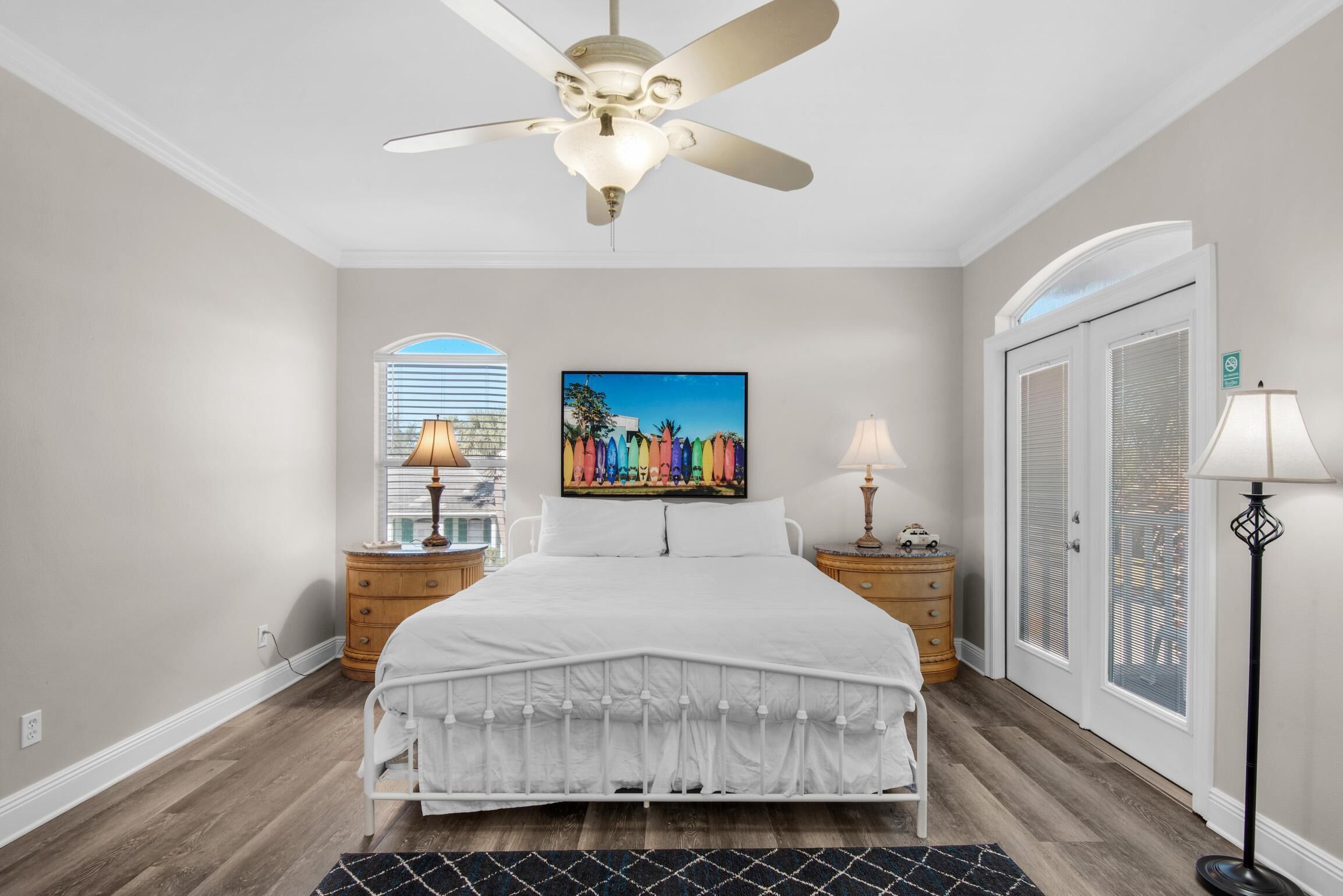A bedroom with a bed, a ceiling fan, two bedside tables with lamps, a window with blinds, and a door with a window.