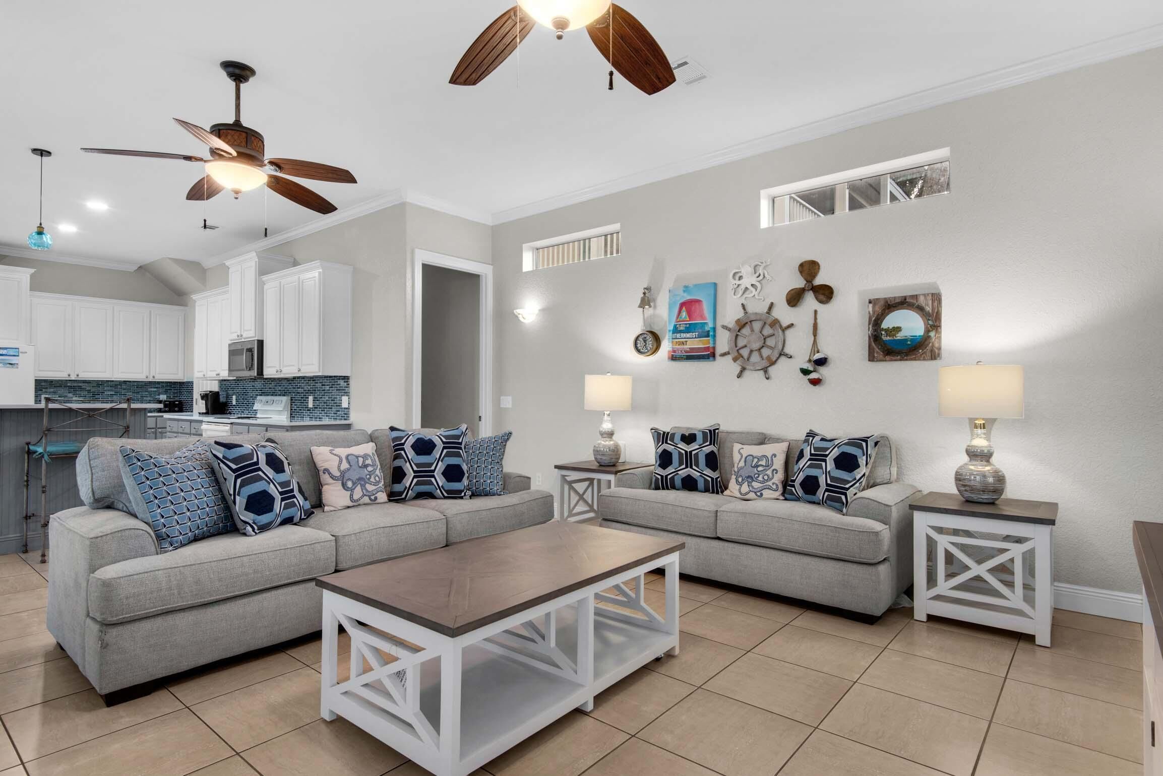 A modern living room with a grey sofa set, a wooden coffee table, and two side tables with lamps. A ceiling fan is visible above the sofa.