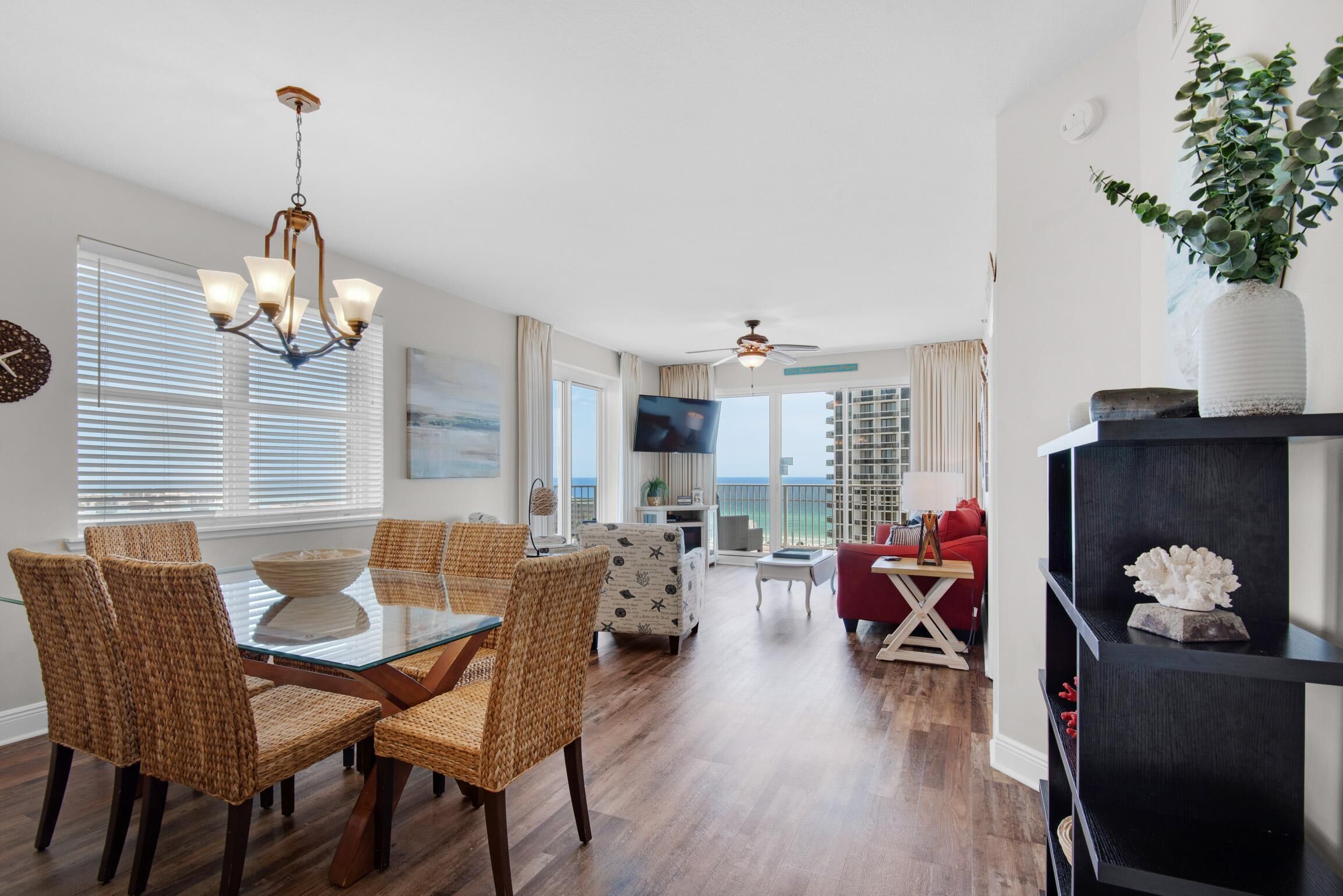 A spacious living area with a dining table, wicker chairs, a ceiling fan, and a large window with blinds.