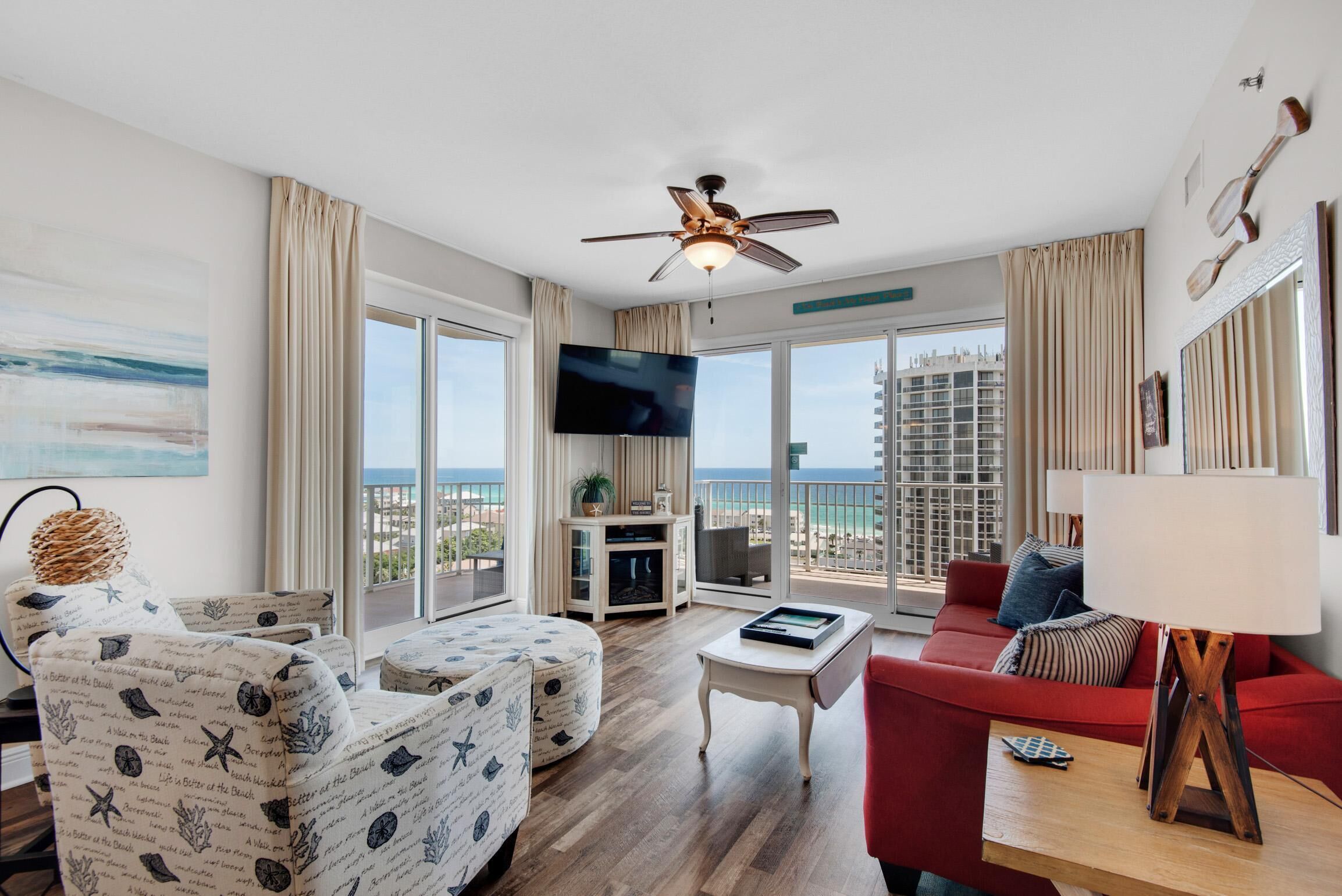 A spacious living room with a red sofa, patterned ottomans, a coffee table, and a television. Large windows offer a view of the city and ocean.